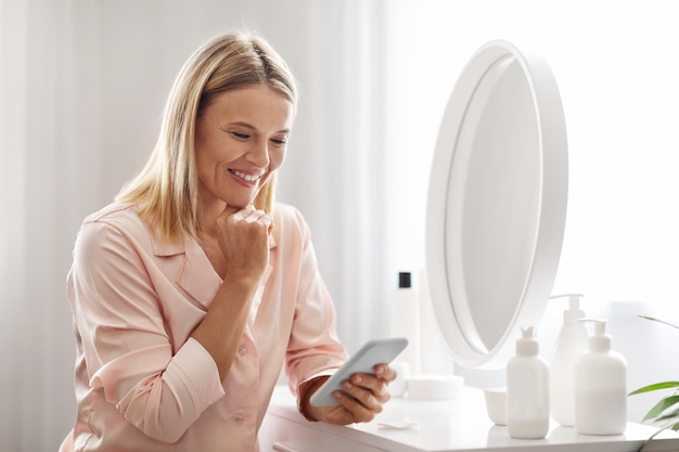 Reife Frau mit Smartphone liest Beauty-Hacks, während sie sich zu Hause fertig macht