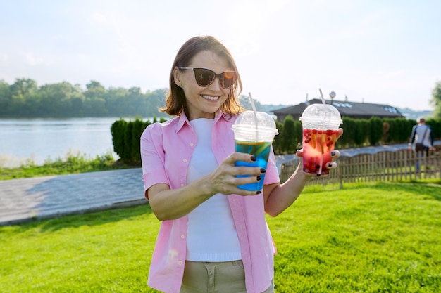 Reife Frau mit zwei Getränken in ihren Händen. Grünes Erholungs- und Unterhaltungsgebiet in der Nähe des Flusses, Kopienraum, goldene Stunde