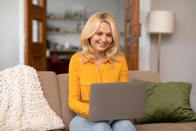 Reife Frau schreibt auf dem Laptop und schaut sich Video im Wohnzimmer an