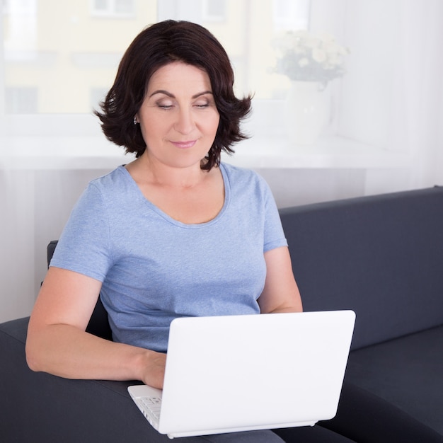 Reife Frau sitzt auf dem Sofa und benutzt den Laptop zu Hause