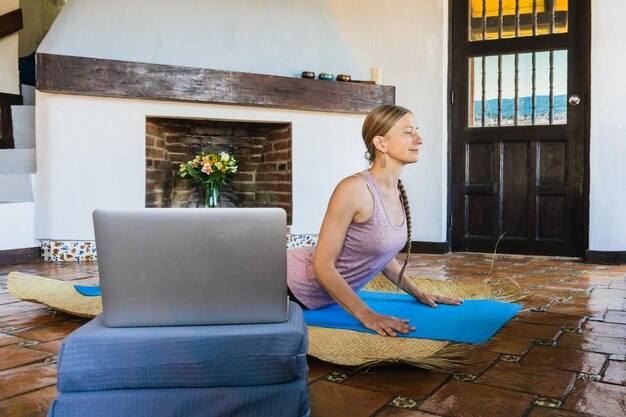 Reife Frau zu Hause, die Yoga hinter einem Computer praktiziert.