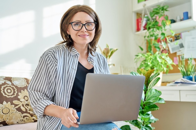 Reife Geschäftsfrau arbeitet auf der Couch mit Laptop und blickt in die Kamera