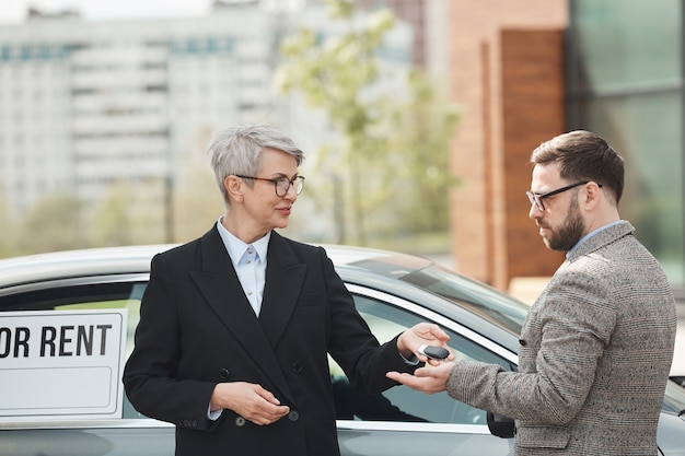 Reife Geschäftsfrau, die die Schlüssel vom Auto zum Geschäftsmann gibt, der sie mietet, die sie draußen stehen