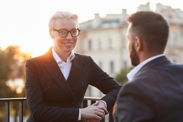 Reife Geschäftsfrau in Brillen und im Anzug lächelnd, während sie mit Geschäftsmann am Balkon während Sonnenuntergang im Hintergrund spricht