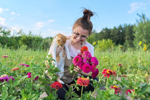 Reife glückliche Frau mit Hauskatze und frischem Zinniablumenblumenstrauß, Sommernaturlandschaftsgemüsegartenhintergrund
