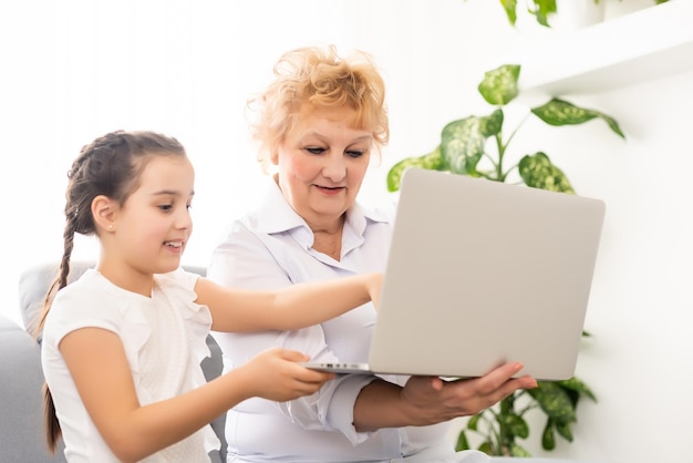 Reife Großmutter hilft Kind bei den Hausaufgaben zu Hause. Zufriedene alte Oma hilft ihrer Enkelin beim Lernen im Wohnzimmer. Kleines Mädchen schreibt auf Notizbuch mit Oberlehrerin, die neben ihr sitzt