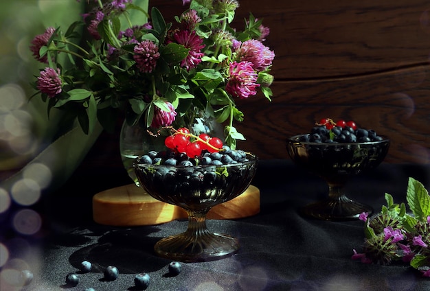 Reife Heidelbeeren in einer Glasschale in der Sonne ein Strauß wilder Blumen in einer Glasvase auf Holzhintergrund Bokeh