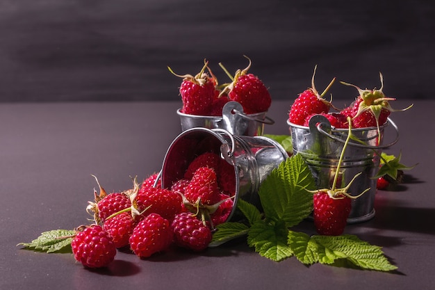 Reife Himbeeren auf schwarzem Steinbetonhintergrund. Kleine Eimer, frische Blätter, neue Ernte. Trendiges hartes Licht, dunkle Schatten, Platz für Text