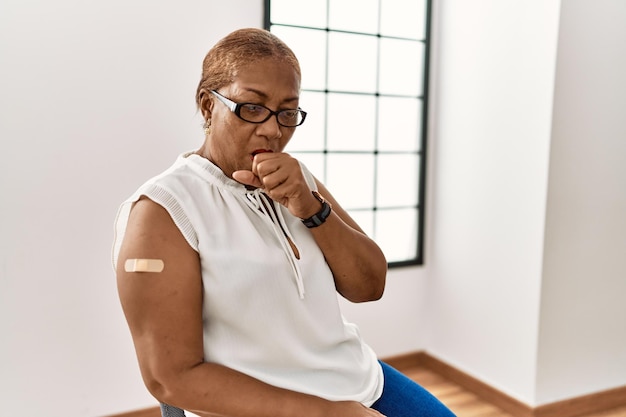 Reife hispanische Frau, die sich impfen lässt, zeigt Arm mit Pflaster, fühlt sich unwohl und hustet als Symptom für Erkältung oder Bronchitis. Gesundheitskonzept