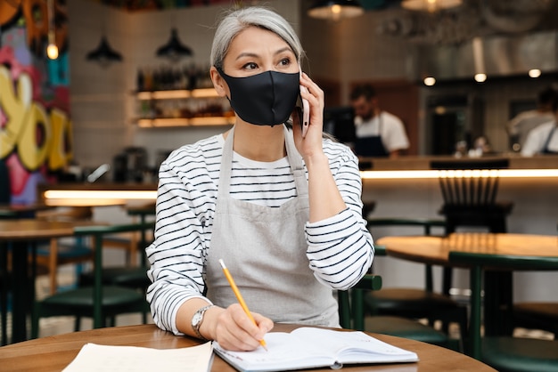 Reife Kellnerin mit Gesichtsmaske, die während der Arbeit am Tisch im Café am Handy spricht