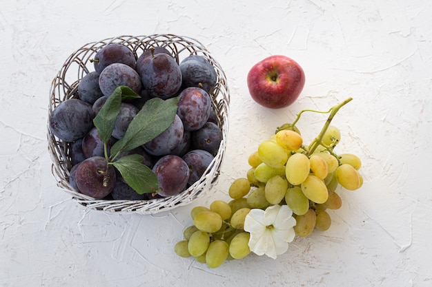 Reife Pflaumen in einem Weidenkorb ein Apfel und ein Bündel reife grüne Trauben auf weißem strukturiertem Hintergrund