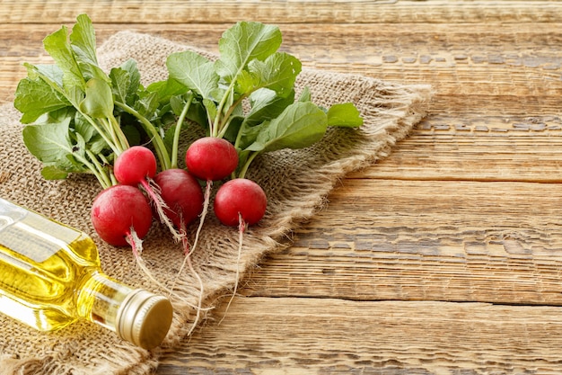 Reife rote Radieschen mit grünen Stielen und einer Flasche Öl auf Sackleinen und alten Holzbrettern. Ansicht von oben. Frühjahrsernte von Gemüse in einem Garten.