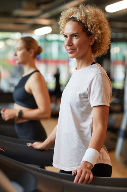 Reife schöne Frau, die auf Laufband im Fitnessstudio mit anderen Leuten im Hintergrund trainiert