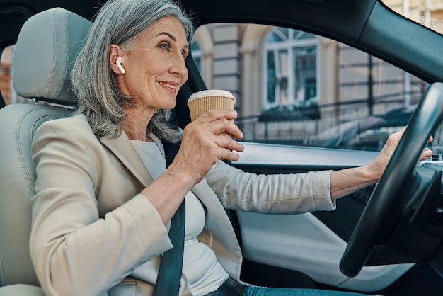 Reife schöne Frau in smart Freizeitkleidung lächelt und trinkt Kaffee beim Autofahren