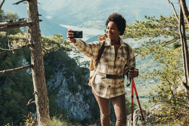 Reife schwarze Frau, die ein Selfie macht, während sie auf einem Hügel steht und die Aussicht während ihrer Wanderung in den Bergen genießt.