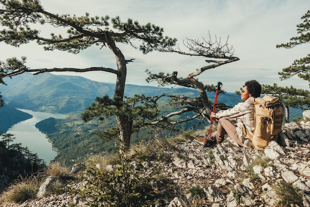 Reife schwarze Frau genießt die Aussicht, während sie während ihres Wanderabenteuers auf einer Bergklippe sitzt.