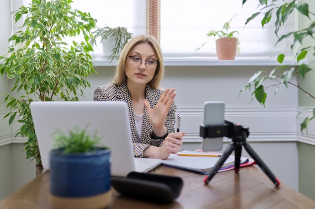 Reife, selbstbewusste Geschäftsfrau, die zu Hause remote arbeitet und Laptop-Smartphones für Videokommunikation und Stream-Aufzeichnung verwendet. Beraterin, Psychologin, Lehrerin, Bloggerin, Anwältin, die online arbeitet