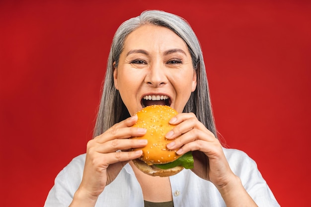 Reife Seniorin isst Burger mit Zufriedenheit Großmutter genießt leckeren Hamburger zum Mitnehmen köstlichen Bissen von Burger bestellen Fastfood-Lieferung, während sie hungrig isoliert auf rotem Hintergrund steht