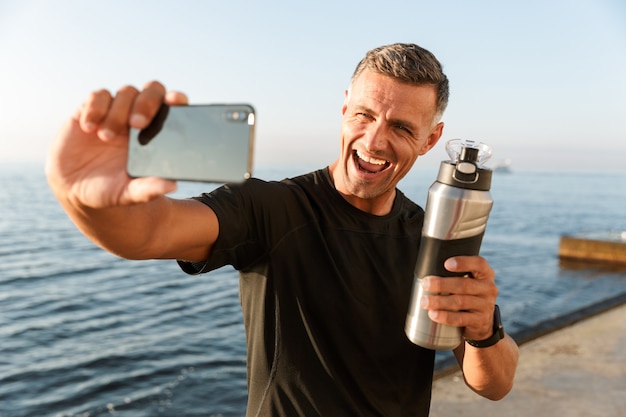 Reife Sportler machen ein Selfie mit einer Flasche Wasser am Strand.