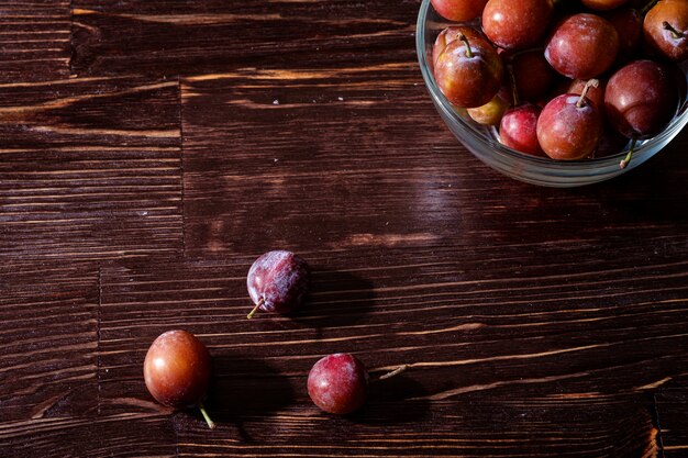 Reife süße Pflaumenfrüchte in Glasschale nahe mit verstreuten Pflaumen auf dunklem launischem Holztisch