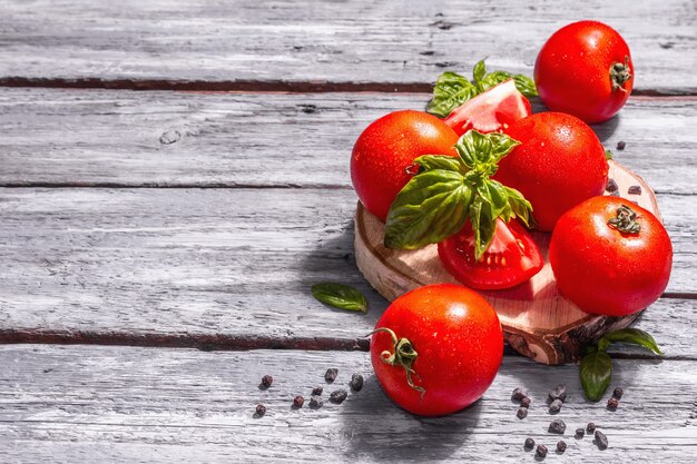 Foto reife tomaten mit frischen basilikumblättern, schwarzem salz und pfefferkörnern. ganzes und halb geschnittenes gemüse, trendiges hartes licht, dunkler schatten. alte holzbretter hintergrund, kopienraum