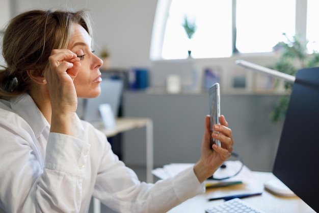 Reife weibliche Büroangestellte, die ihr Make-up repariert, während sie auf die Smartphone-Kamera schauen