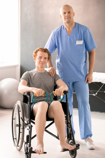 Reifer Arzt in blauer Uniform, der von einem jungen männlichen Patienten im Rollstuhl steht, während beide im medizinischen Zentrum vor der Kamera stehen