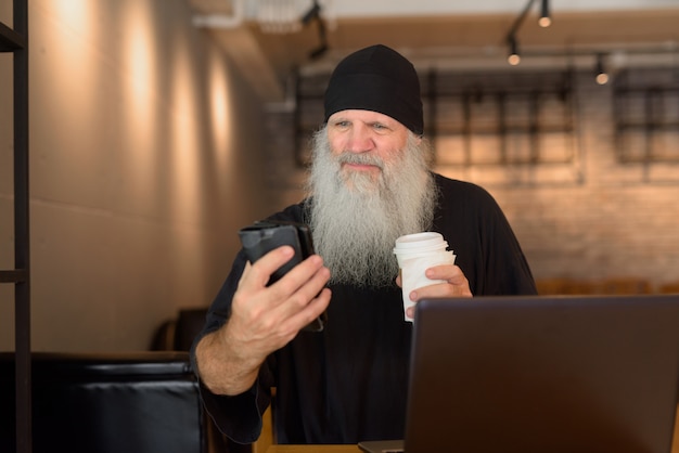 Reifer bärtiger Hipster-Mann, der Telefon benutzt und Kaffee im Kaffeehaus hält