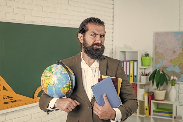 Reifer bärtiger Mann, Lehrer im Kostüm, sitzt im Schulklassenzimmer mit Erdkunde-Globus-Kartenreise
