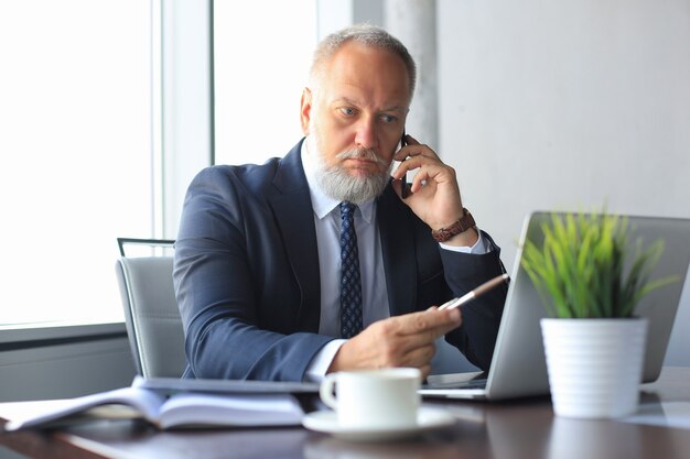 Reifer Geschäftsmann, der am Smartphone spricht und Computer verwendet, während er im Büro arbeitet.