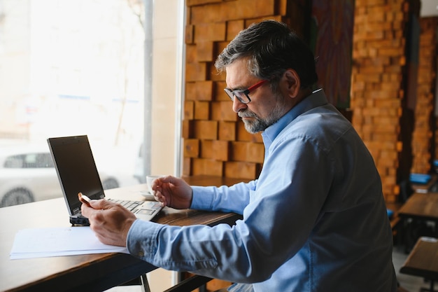 Reifer Geschäftsmann, der Kaffee im Café trinkt Porträt eines gutaussehenden Mannes, der eine stilvolle Brille mit einem Laptop trägt, der lächelnd in die Kamera schaut Kaffeepause-Konzept