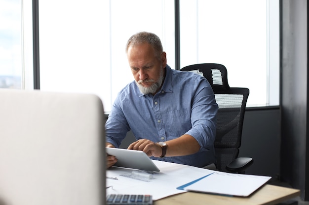 Reifer Geschäftsmann, der mit digitalem Tablet arbeitet, während er im modernen Büro sitzt.