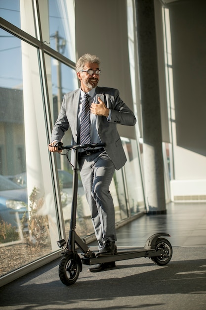 Foto reifer geschäftsmann, der mit elektroroller am bürofenster steht