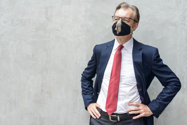 Reifer Geschäftsmann im Anzug denken und Maske gegen Betonwand tragen