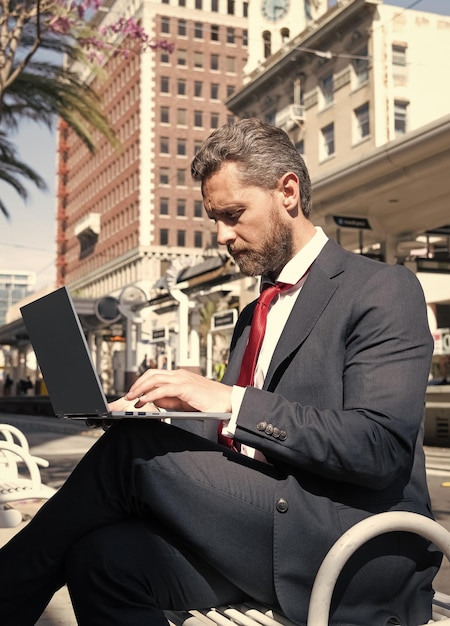 Reifer Geschäftsmann im Anzug sitzt auf einer Bank in der Stadt im Freien und arbeitet online an einem agilen Geschäft mit Laptop