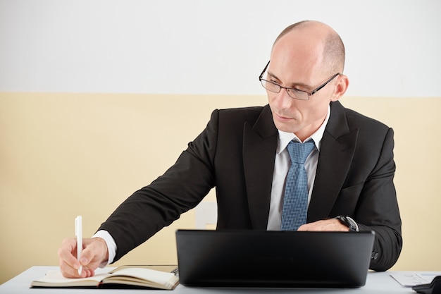 Reifer Geschäftsmann oder Politiker sitzt an seinem Tisch, arbeitet am Laptop und macht sich Notizen im Planer