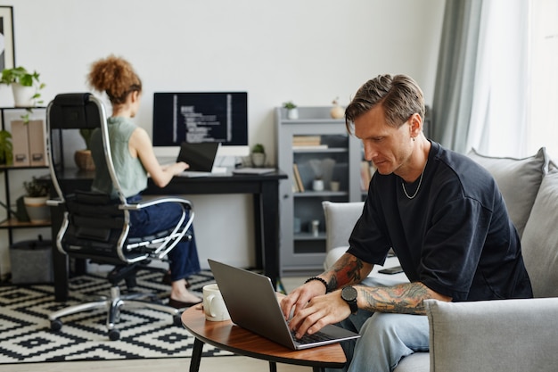 Reifer Geschäftsmann sitzt auf dem Sofa und arbeitet mit seinem Kollegen im Hintergrund am Laptop