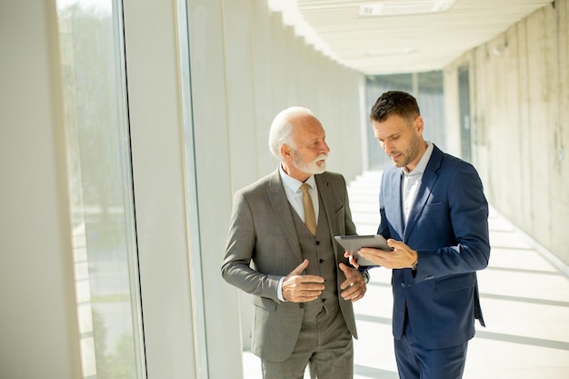 Reifer Geschäftsmann und sein junger Kollege besprechen den Finanzbericht auf dem digitalen Tablet im Bürokorridor