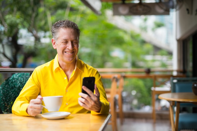 Reifer hübscher Mann, der im Kaffeehaus sitzt, während Handy benutzt