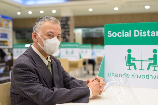 Reifer japanischer Geschäftsmann, der Maske trägt und mit Abstand am Food Court sitzt