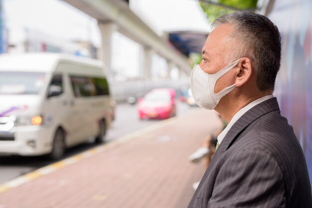 Reifer japanischer Geschäftsmann mit Maske, die mit Abstand an der Bushaltestelle sitzt