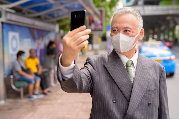 Reifer japanischer Geschäftsmann mit Maske, die Selfie an der Bushaltestelle nimmt