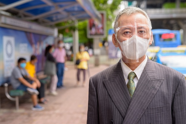 Reifer japanischer Geschäftsmann mit Maske und Gesichtsschutz an der Bushaltestelle