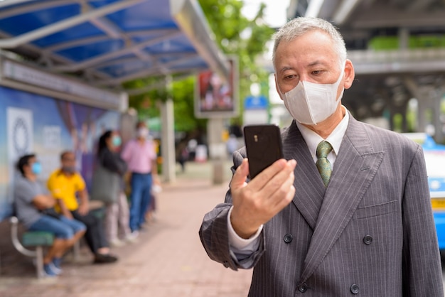 Reifer japanischer Geschäftsmann mit Maske unter Verwendung des Telefons an der Bushaltestelle