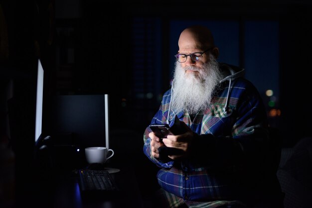 Reifer kahler bärtiger Hipster-Mann, der spät in der Nacht im Dunkeln zu Hause telefoniert