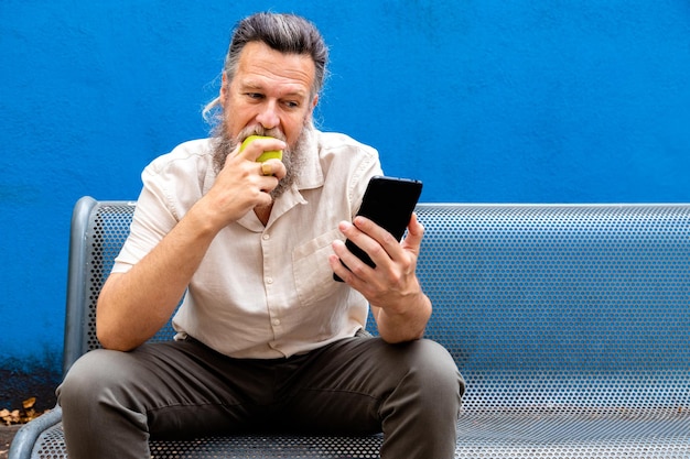 Reifer kaukasischer Mann, der Handy benutzt und einen Apfel in einem öffentlichen Park isst Kopieren Sie Platz Lifestyle