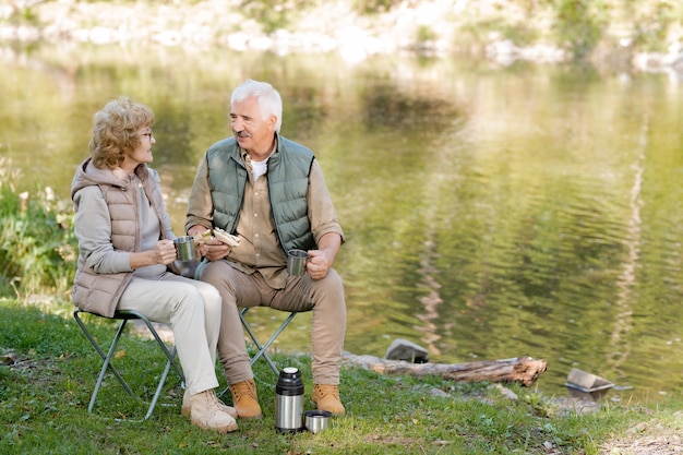 Reifer liebevoller Mann und Frau in Aktivkleidung, die Tee mit Sandwiches beim Sitzen auf Touristenstühlen am Fluss trinken