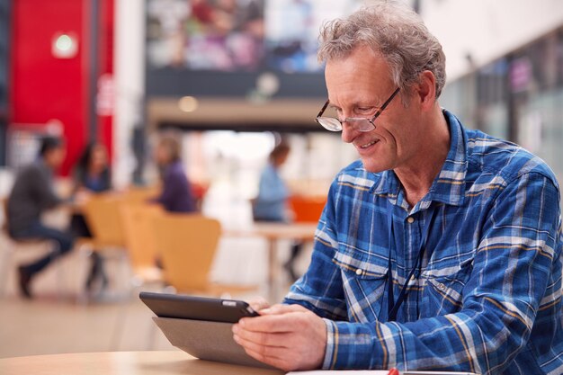 Reifer männlicher Lehrer oder Student mit digitalem Tablet, der am Tisch in der College-Halle arbeitet