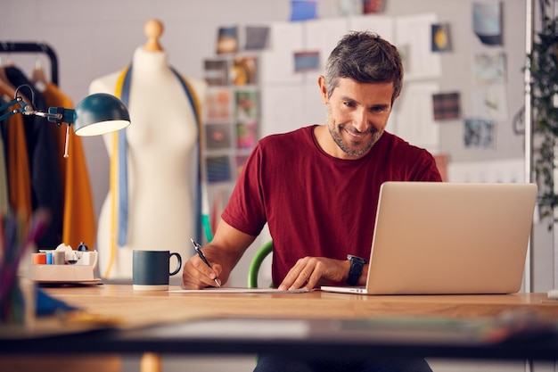 Reifer männlicher Modedesigner im Studio, der an Skizzen oder Dokumenten am Schreibtisch mit Laptop arbeitet