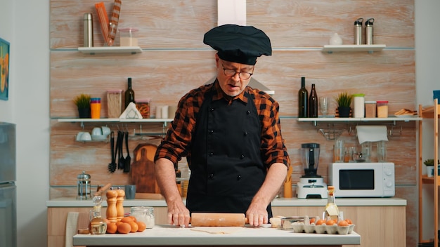 Reifer Mann, der hausgemachte Pizza zu Hause zubereitet. Glücklicher älterer Koch mit Knochen, der mit Holznudelholz rohe Zutaten knetet, um traditionelle Kekse zu backen, Mehl auf dem Tisch zu bestreuen und zu sieben.